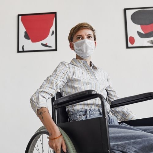 A person with a mask sitting on a wheel chair with two pieces of artwork on a wall behind them